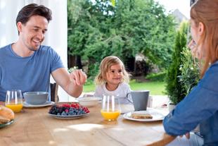 Eine dreiköpfige Familie, bestehend aus einem Mann, einer Frau und einem jungen Mädchen, genießt ein Frühstück im Freien mit frischem Obst und Getränken.