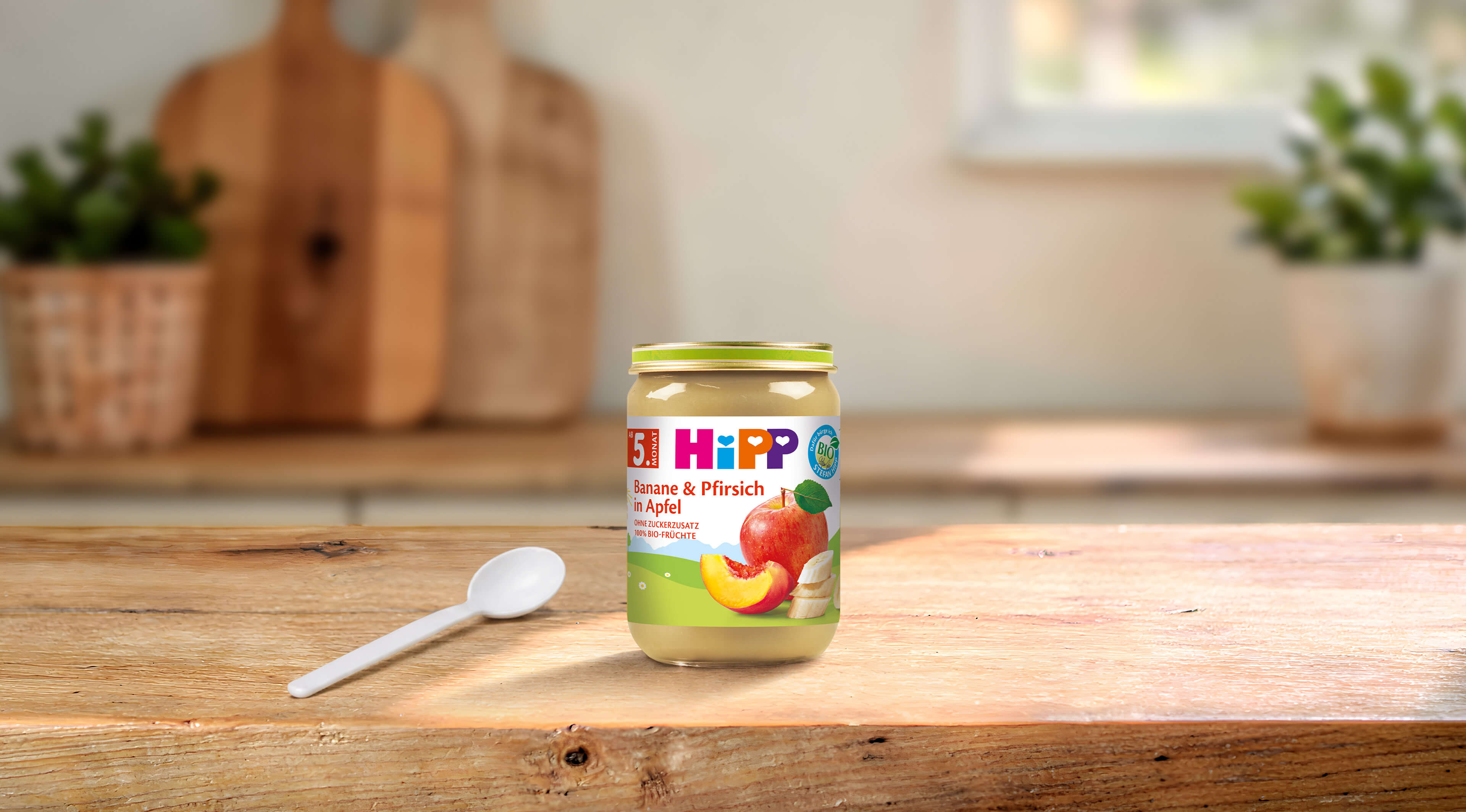 HiPP Bio Babygläschen „Banane & Pfirsich in Apfel“ ab 5. Monat mit Löffel auf Holztisch in heller Küche