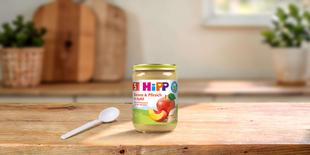 HiPP Bio Babygläschen „Banane & Pfirsich in Apfel“ ab 5. Monat mit Löffel auf Holztisch in heller Küche