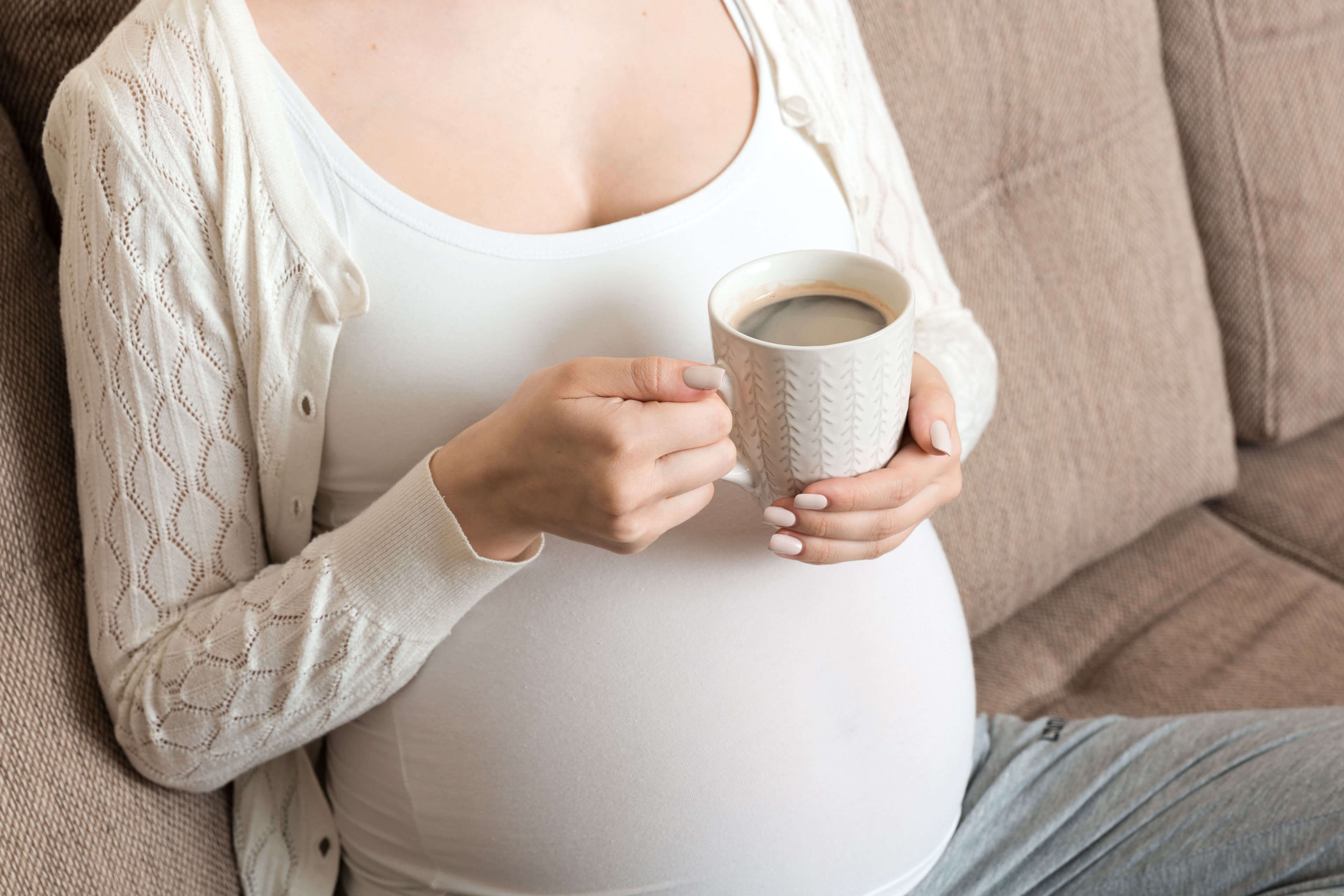 Eine Person mit einer Tasse Kaffee in der Hand, die ein weißes Oberteil und eine Strickjacke trägt, auf der ein schwangerer Bauch zu sehen ist.