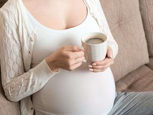 Eine Person mit einer Tasse Kaffee in der Hand, die ein weißes Oberteil und eine Strickjacke trägt, auf der ein schwangerer Bauch zu sehen ist.