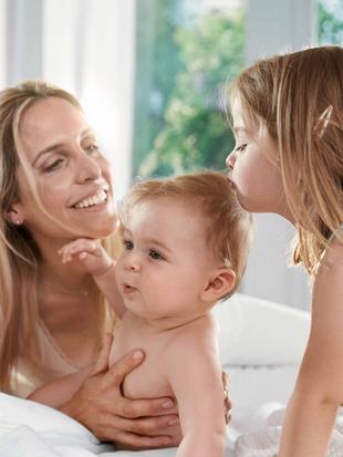 Ein freudiger Moment zwischen einer Mutter und ihren beiden Kindern, wobei die Mutter ein Baby im Arm hält und ein junges Mädchen sich vorbeugt, um das Baby zu küssen.
