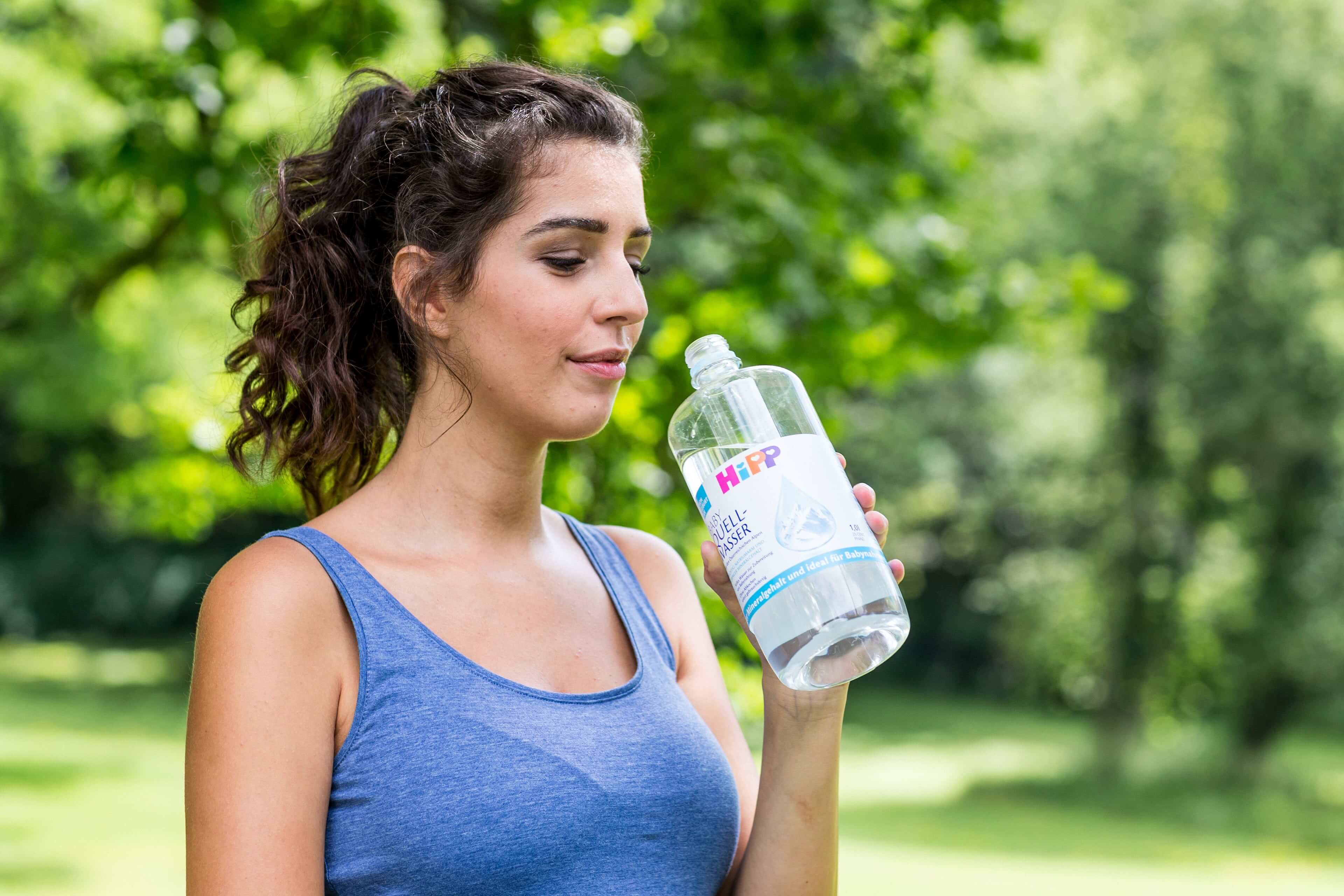 Eine Frau in einem blauen Tank-Top trinkt Wasser aus einer durchsichtigen Flasche mit der Aufschrift "Nature's Pure".
