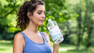 Eine Frau in einem blauen Tank-Top trinkt Wasser aus einer durchsichtigen Flasche mit der Aufschrift "Nature's Pure".
