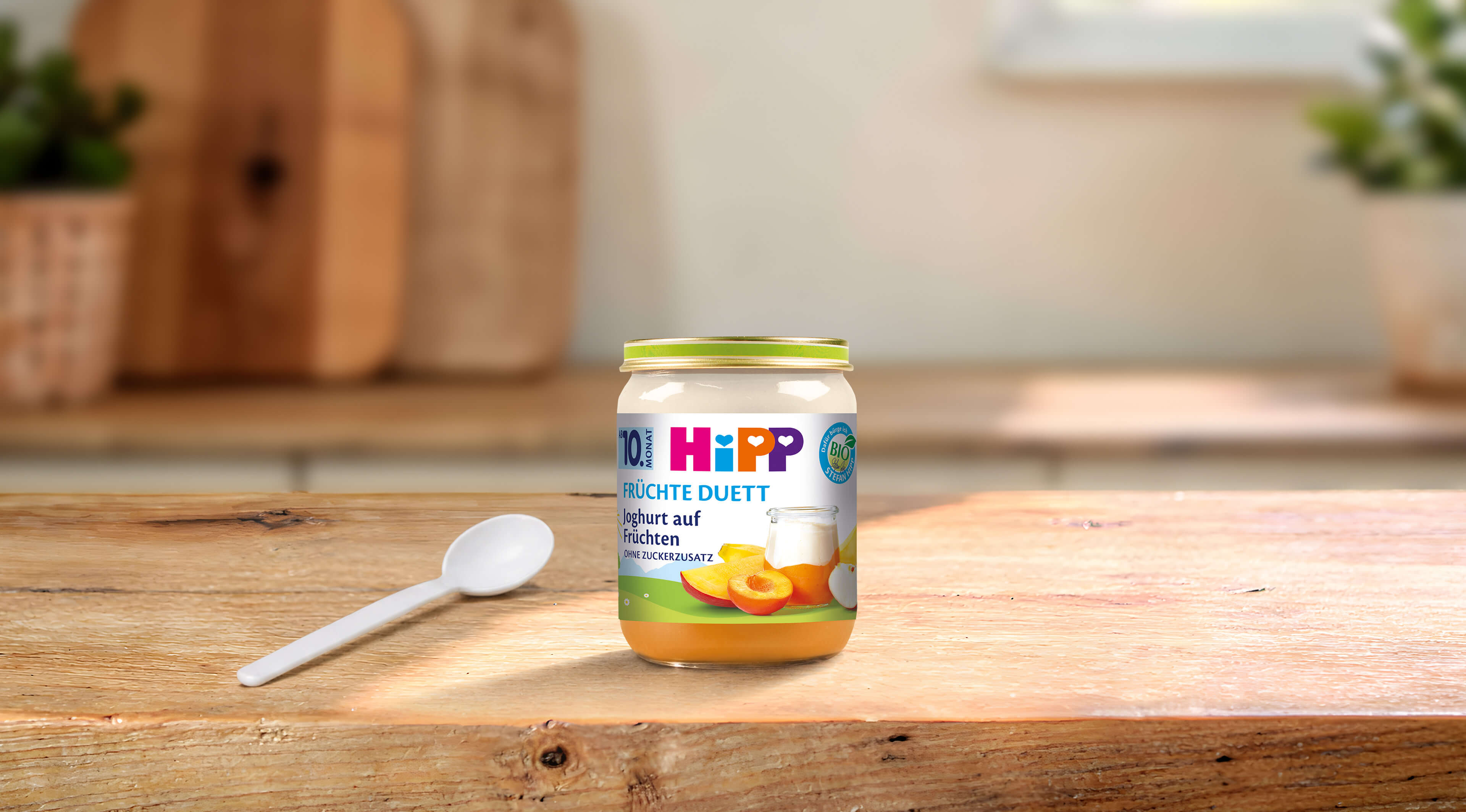 HiPP Früchte Duett Joghurt auf Früchten im Glas mit Aprikosenmotiv und Löffel auf Holztisch in heller Küche