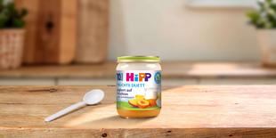 HiPP Früchte Duett Joghurt auf Früchten im Glas mit Aprikosenmotiv und Löffel auf Holztisch in heller Küche