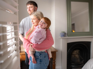 Toddler smiling at camera whilst mum and dad look out the window