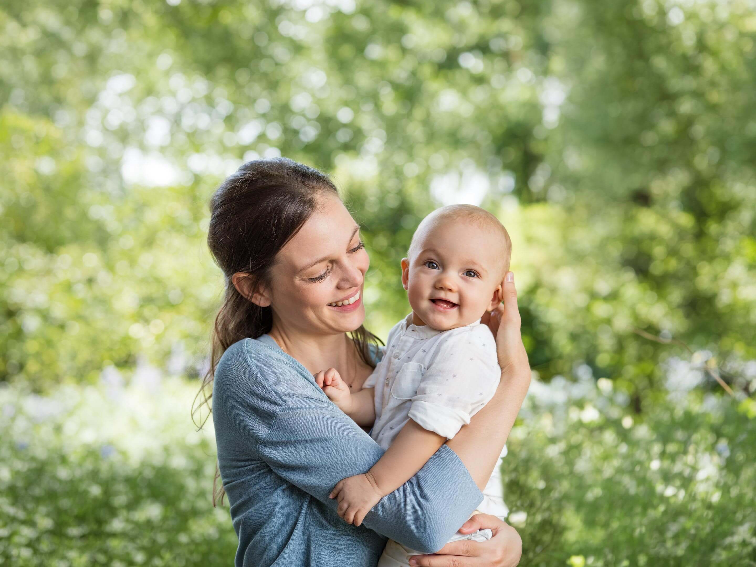 Lächelnde Mutter hält ihr fröhliches Baby im Grünen, umgeben von natürlicher, heller Gartenkulisse