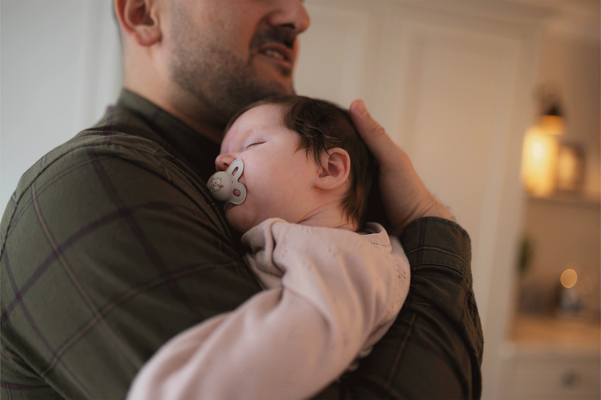 A father lovingly holds his sleeping newborn baby, who has a pacifier in her mouth, resting against his chest.