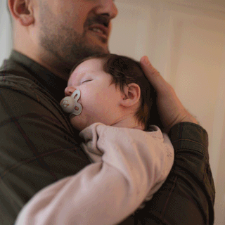 A father lovingly holds his sleeping newborn baby, who has a pacifier in her mouth, resting against his chest.