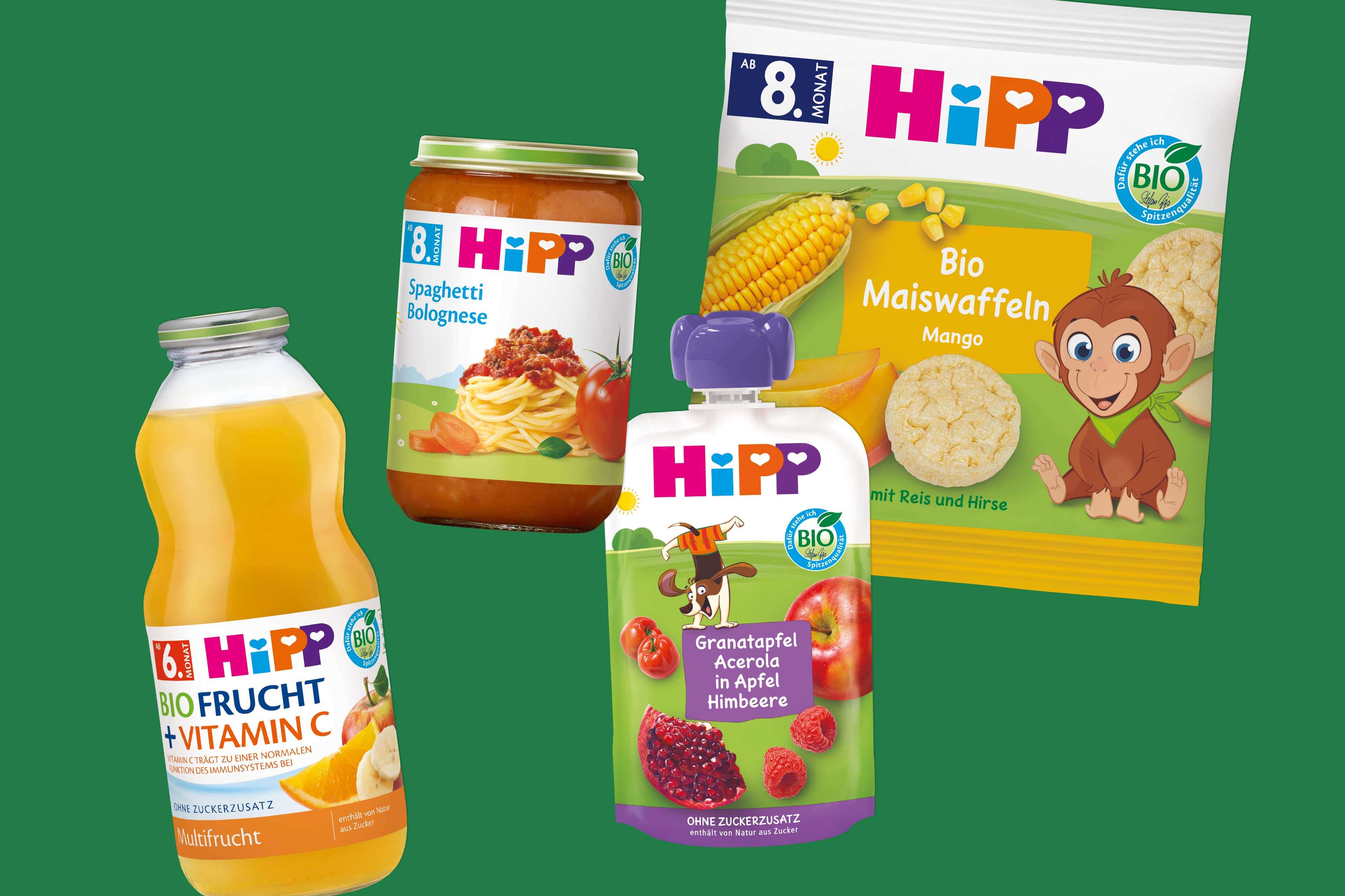 Vier HiPP-Bio-Kinderprodukte: Fruchtsaft mit Vitamin C, Spaghetti Bolognese im Glas, Fruchtpüree im Quetschbeutel und Bio-Maiswaffeln mit Mango, vor grünem Hintergrund