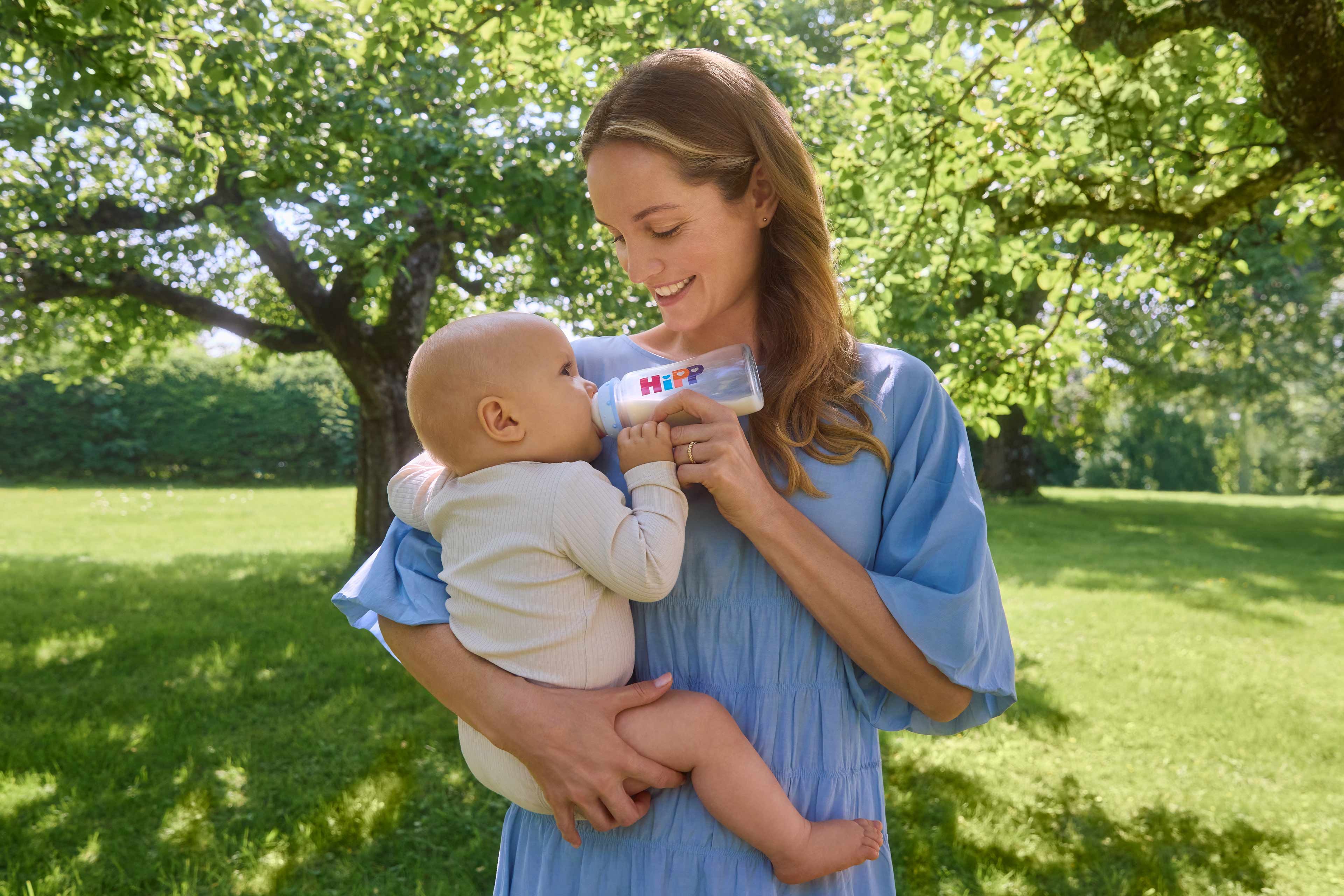 Frau in hellblauem Kleid hält ein Baby im Arm und füttert es mit einer HiPP-Babymilchflasche im Garten
