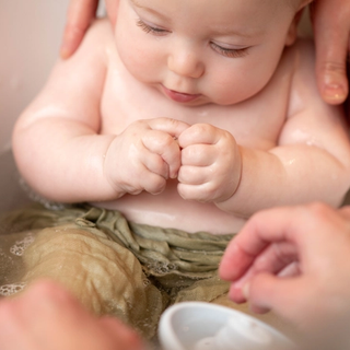 A close-up of a baby taking a bath, sitting in a small tub with their hands clenched together. The baby is partially submerged in water, wearing light green cloth around the waist. Two adult hands are gently supporting and washing the baby.
