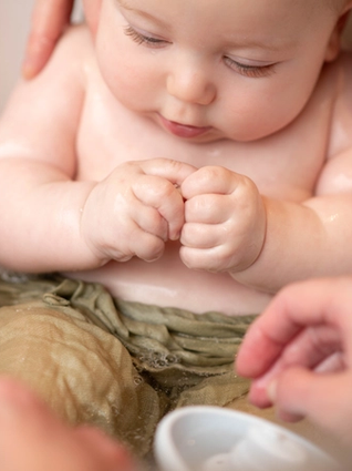 A close-up of a baby taking a bath, sitting in a small tub with their hands clenched together. The baby is partially submerged in water, wearing light green cloth around the waist. Two adult hands are gently supporting and washing the baby.