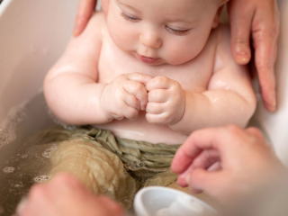 A close-up of a baby taking a bath, sitting in a small tub with their hands clenched together. The baby is partially submerged in water, wearing light green cloth around the waist. Two adult hands are gently supporting and washing the baby.