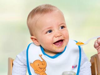 Lachendes Baby mit Lätzchen wird liebevoll mit Beikost vom Löffel gefüttert
