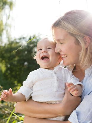 Ein zärtlicher Moment zwischen einer Mutter und ihrem Baby, wobei die Mutter ihr Kind im Arm hält und beide einen freudigen Moment inmitten einer natürlichen Kulisse erleben.