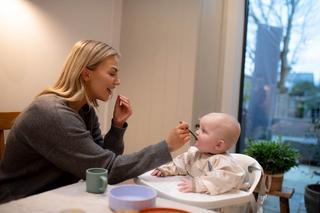 A woman smiles while feeding a baby sitting in a high chair, gently offering a spoonful of food as the baby looks up attentively. A cozy indoor setting with a view of the backyard adds warmth to this mealtime moment