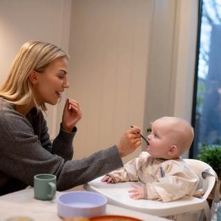 A woman smiles while feeding a baby sitting in a high chair, gently offering a spoonful of food as the baby looks up attentively. A cozy indoor setting with a view of the backyard adds warmth to this mealtime moment.
