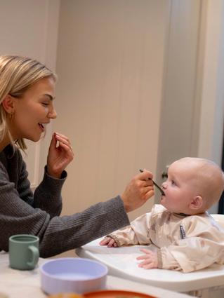 A woman smiles while feeding a baby sitting in a high chair, gently offering a spoonful of food as the baby looks up attentively. A cozy indoor setting with a view of the backyard adds warmth to this mealtime moment.