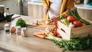 Eine schwangere Frau in einer Küche, die Knoblauch auf einem Holzbrett hackt, umgeben von frischem Gemüse und Gewürzen.
