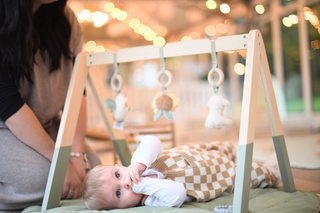 A baby lies on a cushioned mat beneath a wooden play gym, sucking their thumb while gazing up at the hanging plush toys shaped like animals. A woman nearby watches lovingly, surrounded by cozy lighting and a warm, softly lit indoor setting.