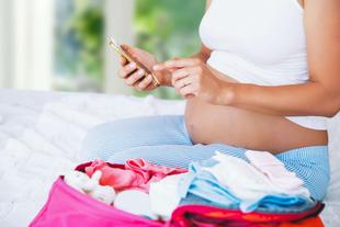 Eine schwangere Frau sitzt auf einem Bett, hält ein Mobiltelefon in der Hand und hat eine rosa Tasche mit Babyartikeln dabei.