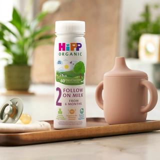HiPP Organic Follow On Milk on a wooden tray next to a beaker and dummy