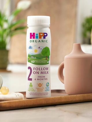 HiPP Organic Follow On Milk on a wooden tray next to a beaker and dummy
