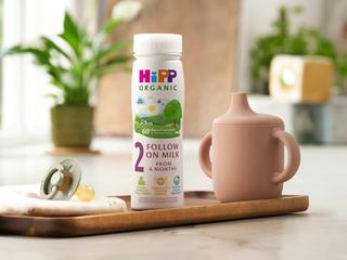 HiPP Organic Follow On Milk on a wooden tray next to a beaker and dummy