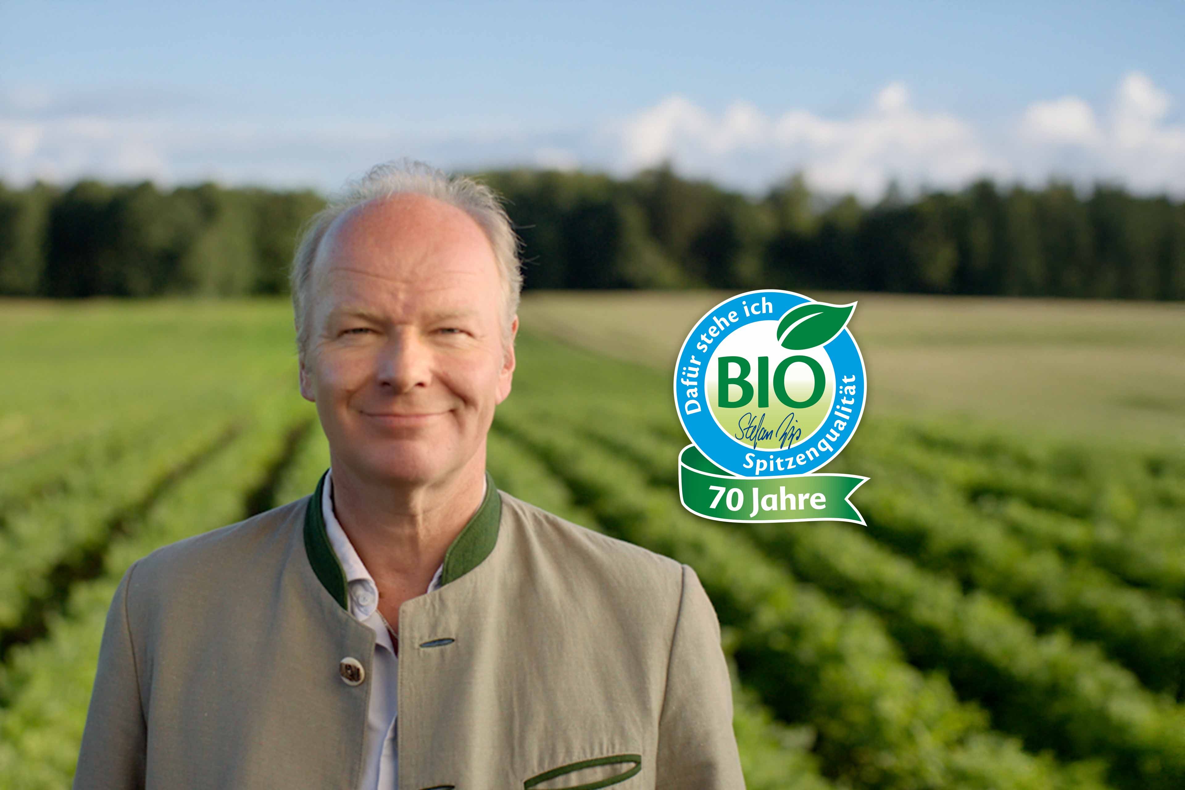 Stefan Hipp in traditioneller Trachtenjacke steht vor einem Feld mit Reihen von Pflanzen und einem Bio-Siegel mit dem Text '70 Jahre'
