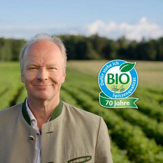 Älterer Mann auf grüner Bio-Ackerlandschaft mit HiPP Bio-Siegel „Dafür stehe ich – 70 Jahre Spitzenqualität“ im Vordergrund.