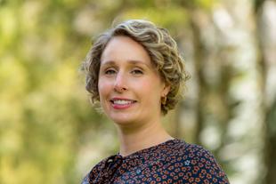 Eine Frau mit lockigem blondem Haar, die eine Bluse mit Blumenmuster trägt, vor einem unscharfen Hintergrund mit Bäumen.