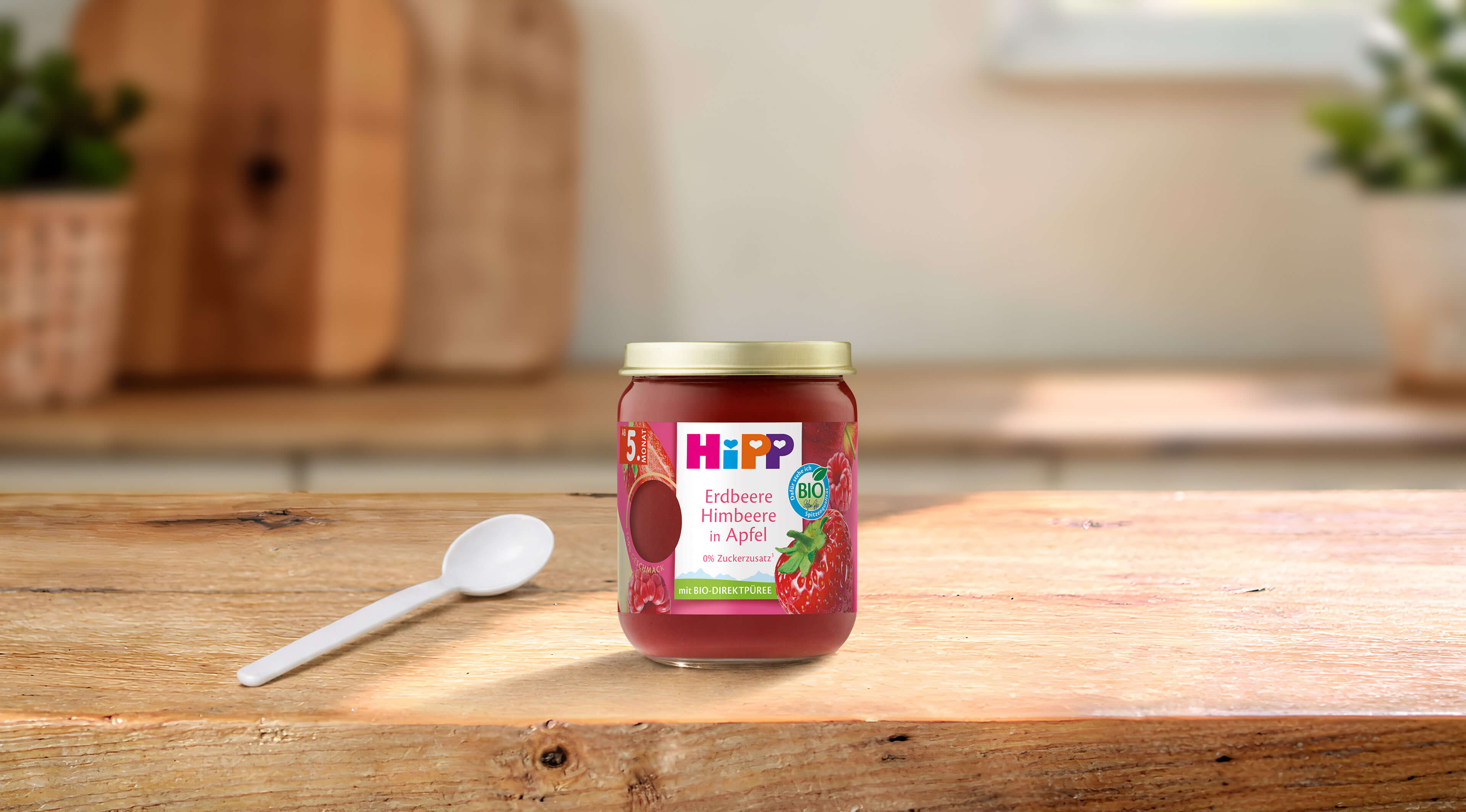HiPP Bio-Früchte-Gläschen Erdbeere Himbeere in Apfel mit Löffel auf Holztisch in heller, gemütlicher Küche