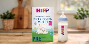 HiPP PRE Anfangsmilch aus Bio Ziegenmilch Packung mit Ziege und Zicklein neben gefülltem Fläschchen auf Holztisch in Küche