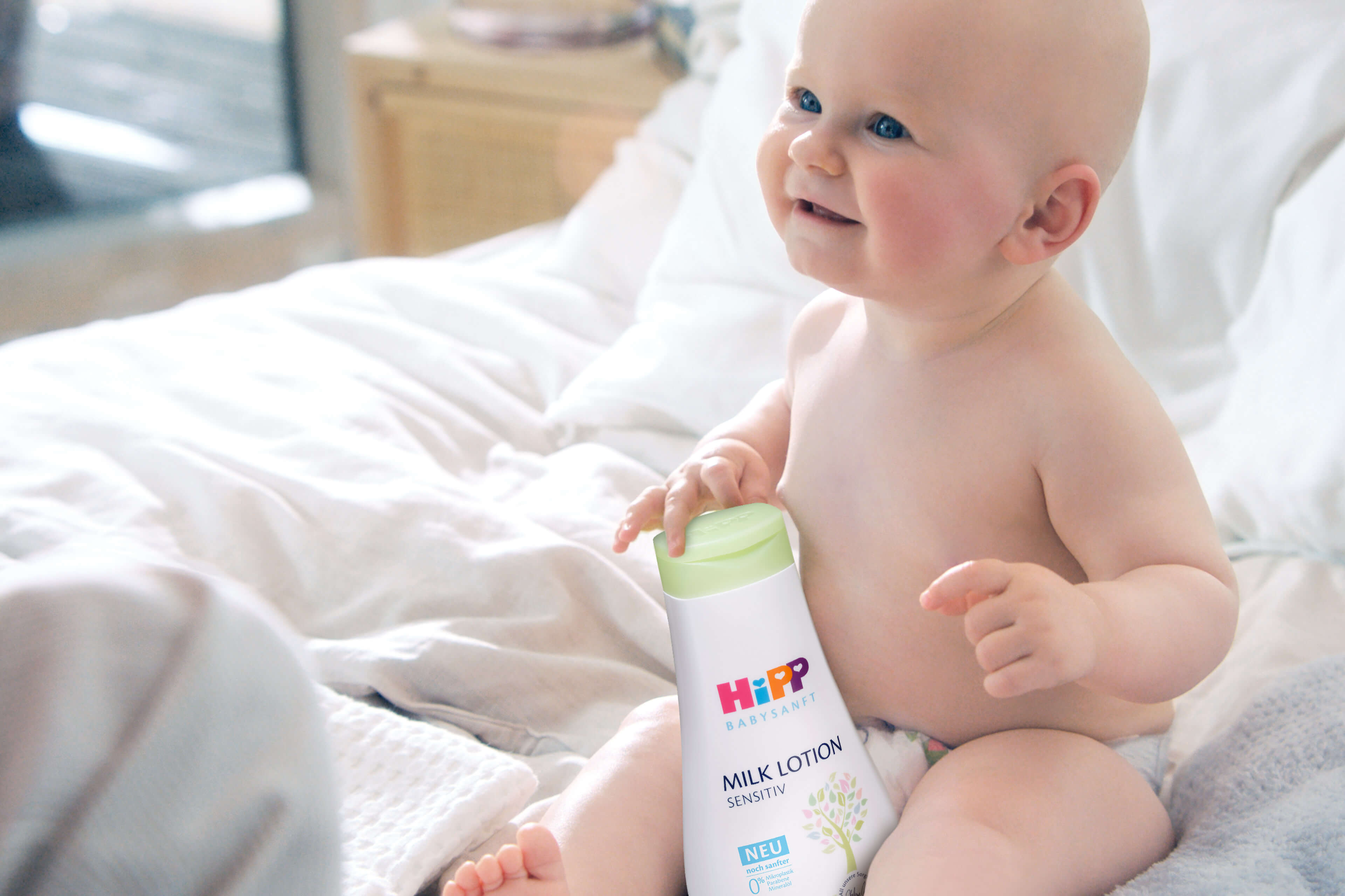 Ein Baby, das auf einem Bett sitzt und eine Flasche mit der Aufschrift "HiPP Milk Lotion Sensitive" in der Hand hält, vor dem Hintergrund eines Zimmers und eines Fensters.