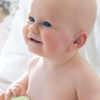 Ein Baby, das auf einem Bett sitzt und eine Flasche mit der Aufschrift "HiPP Milk Lotion Sensitive" in der Hand hält, vor dem Hintergrund eines Zimmers und eines Fensters.