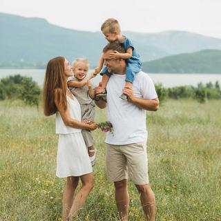 Familie mit zwei Kindern steht auf einer Wiese vor einem See und Bergen im Hintergrund