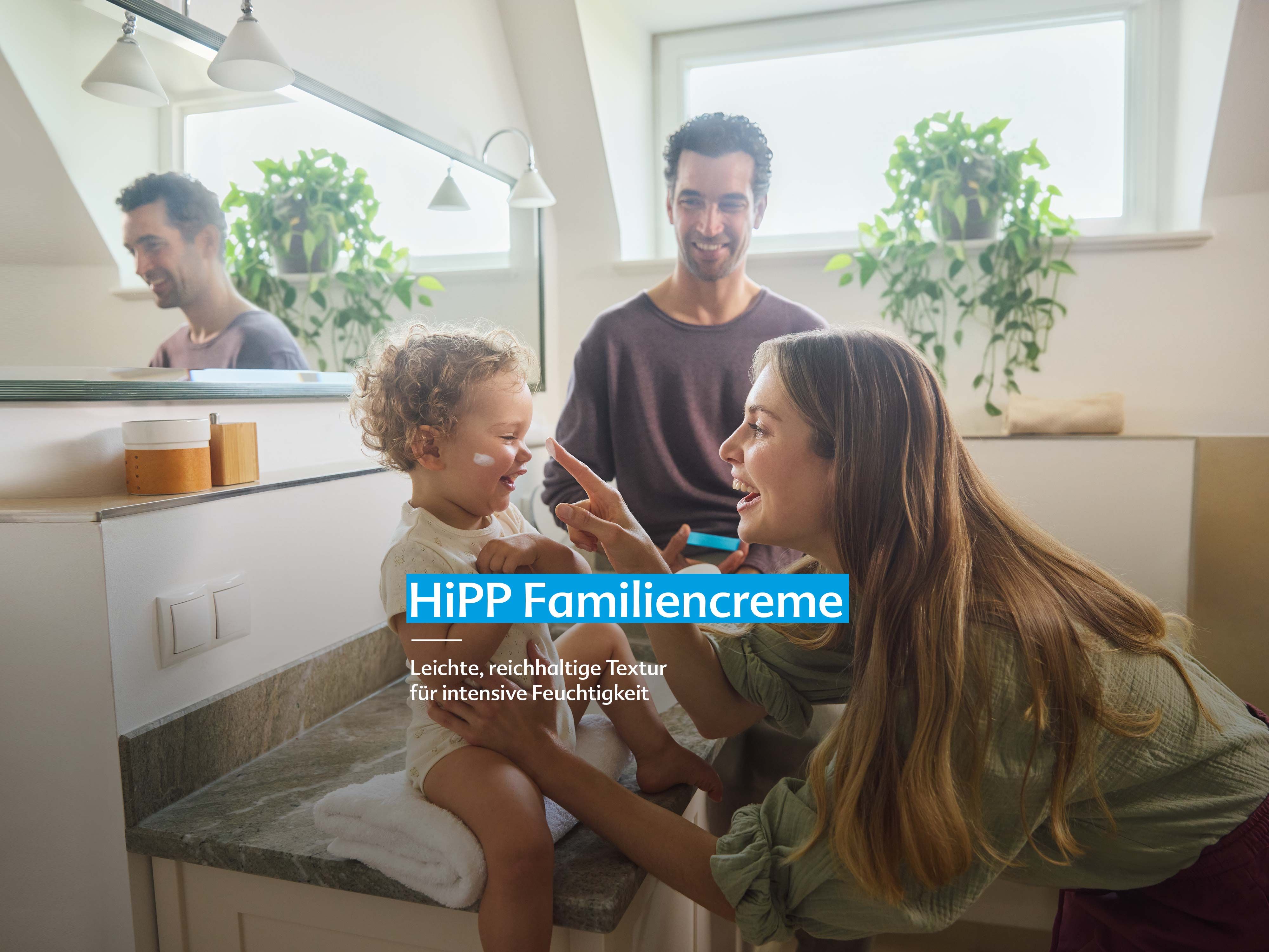Mutter trägt einem lachenden Kleinkind im Badezimmer Creme auf, im Spiegel ist ein Mann zu sehen