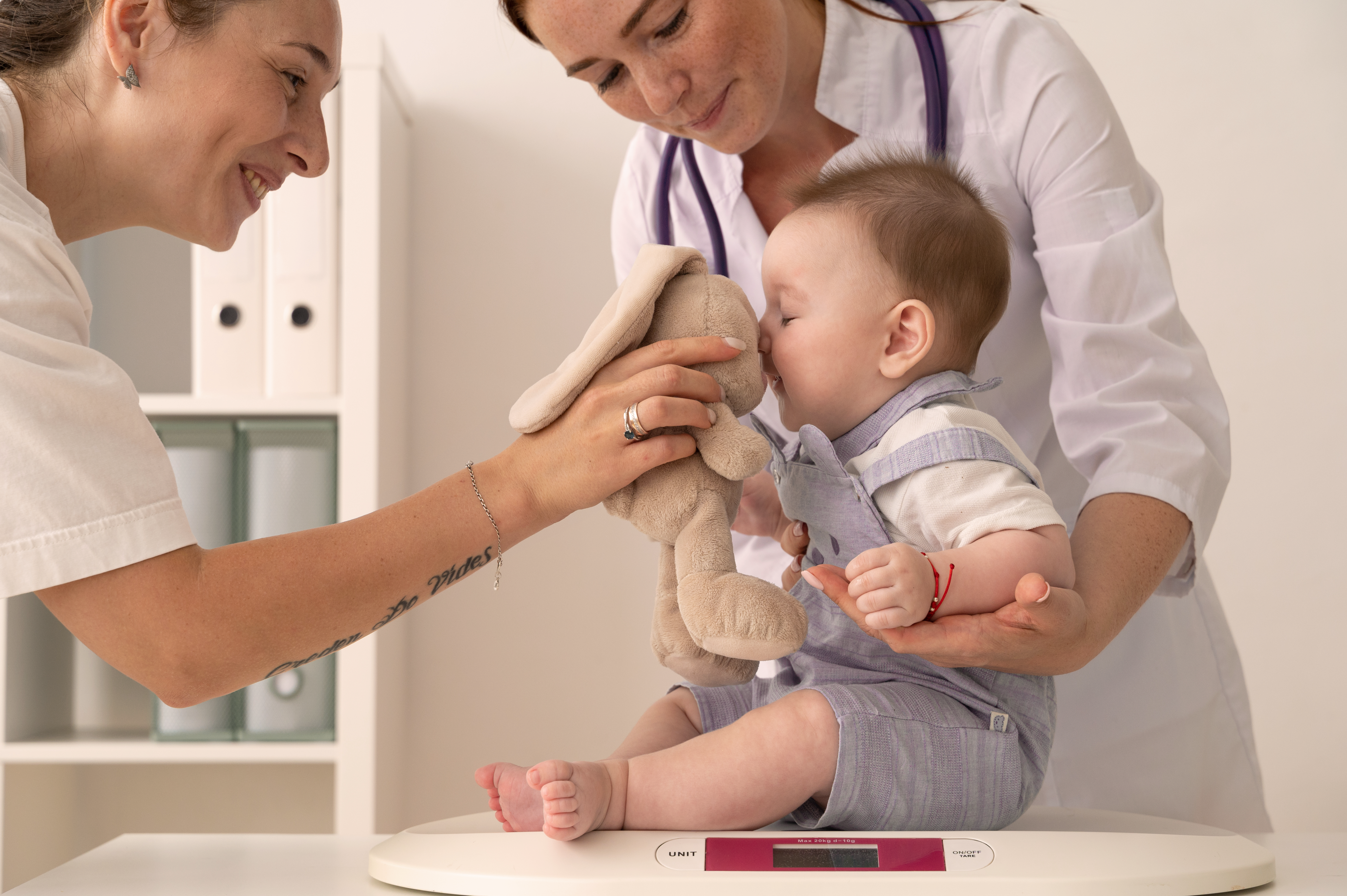 Smiling healthcare professionals gently weigh a baby on scales while comforting them with a soft toy.