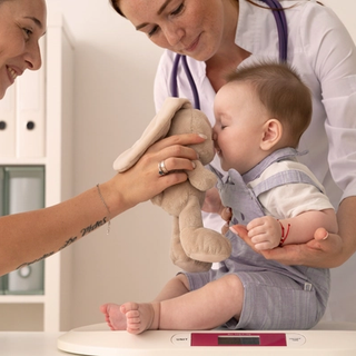 Smiling healthcare professionals gently weigh a baby on scales while comforting them with a soft toy.