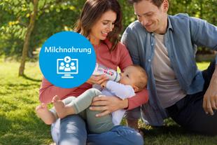 Frau sitzt auf dem Rasen und hält ein Baby im Arm, das aus einer Flasche trinkt, daneben sitzt ein Mann, vorn ein runder blauer Kreis mit dem Wort 'Milchnahrung' und einem Symbol.