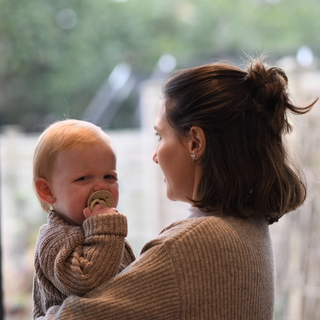 A woman holding a baby who appears to be upset or crying, with a pacifier in their mouth. Both are dressed in warm, knitted sweaters. The woman is facing away from the camera while the baby looks toward it. They are standing indoors near a large window with a blurred view of a garden or backyard in the background.