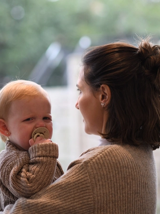 A woman holding a baby who appears to be upset or crying, with a pacifier in their mouth. Both are dressed in warm, knitted sweaters. The woman is facing away from the camera while the baby looks toward it. They are standing indoors near a large window with a blurred view of a garden or backyard in the background.