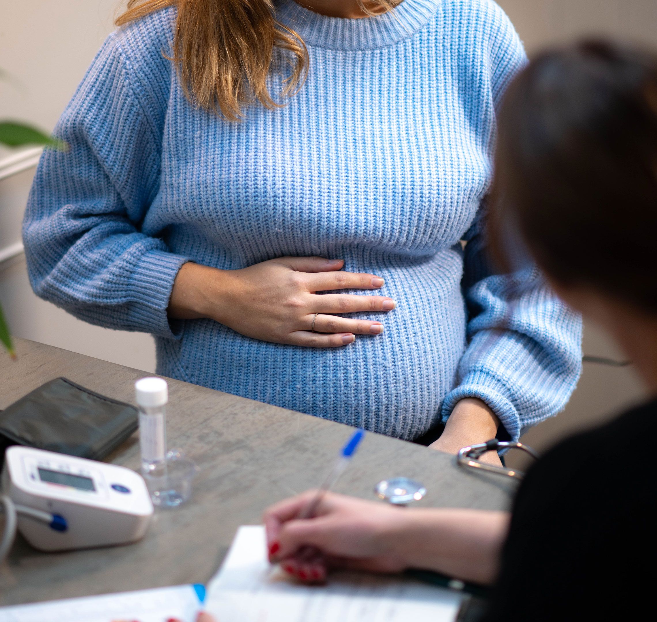 Pregnant woman in a blue jumper holding her bump while talking to a healthcare professional at a check-up.