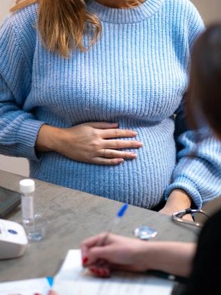 Pregnant woman in a blue jumper holding her bump while talking to a healthcare professional at a check-up.