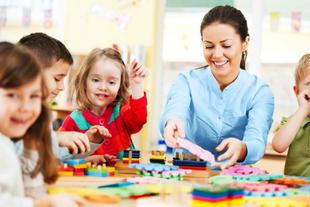 Eine fröhliche Szene in einem Klassenzimmer, in dem ein Lehrer und drei kleine Kinder spielerisch mit bunten Bauklötzen beschäftigt sind.