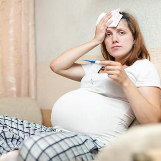 Eine Frau mit einem Thermometer in der Hand, die sich ein Tuch auf die Stirn hält und besorgt dreinschaut.