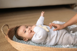 Smiling baby lying in a cosy basket, gently touched by a parent’s hand, suggesting calm, happy family time.