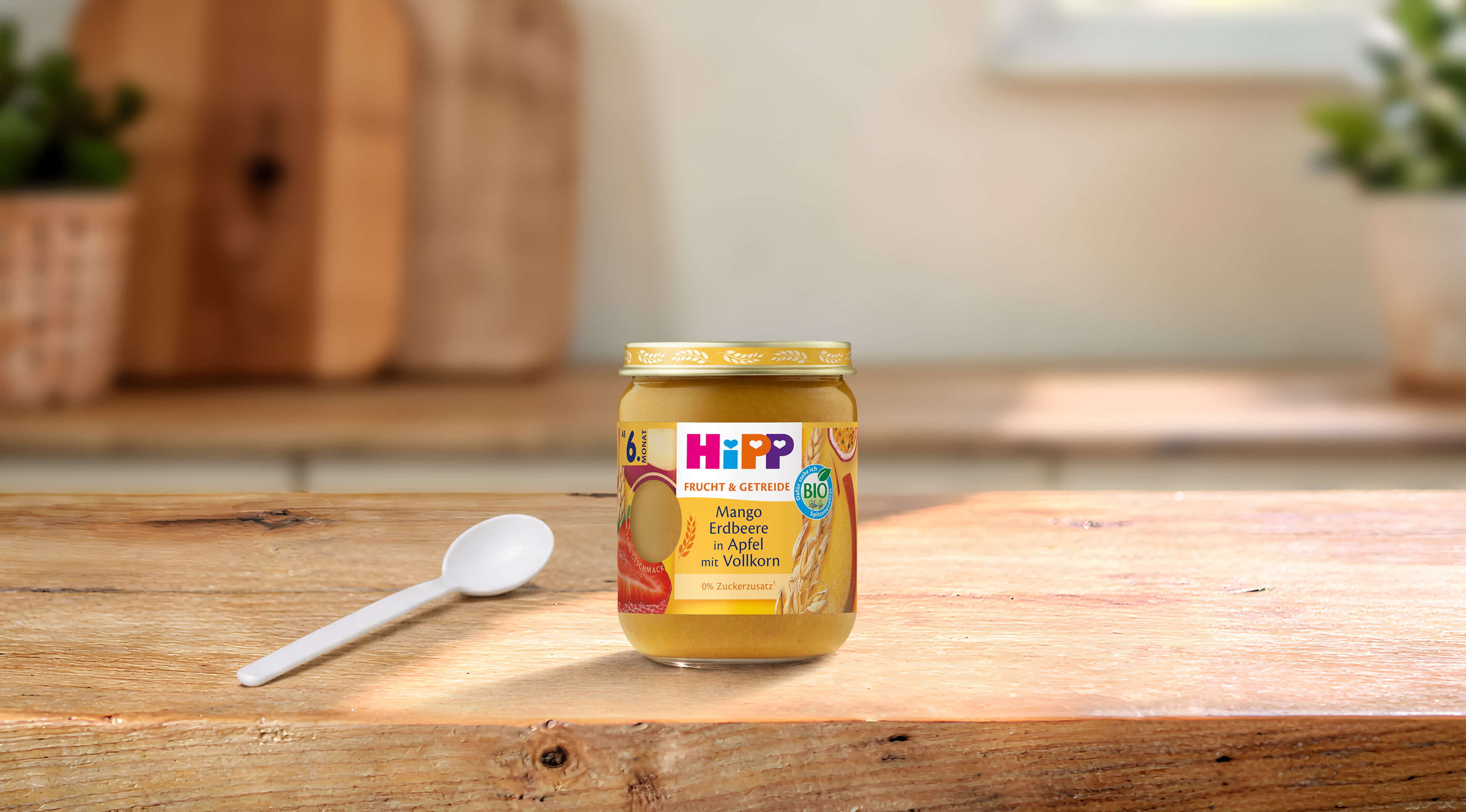 HiPP Bio-Gläschen Mango Erdbeere in Apfel mit Vollkorn auf Holztisch, daneben ein Löffel in heller Küchenumgebung
